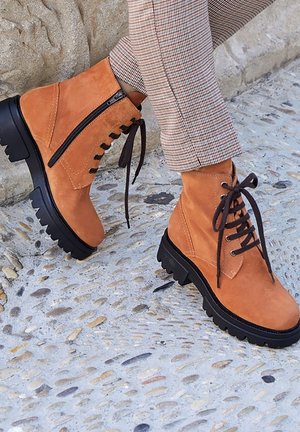 Botines de ante en color tostado con diseño de cordones en la parte delantera, cremallera lateral y suela de goma negra texturizada con un perfil robusto.