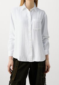 Chemise blanche à manches longues avec boutons, confectionnée en tissu texturé, dotée d'une poche poitrine et d'un ourlet incurvé.