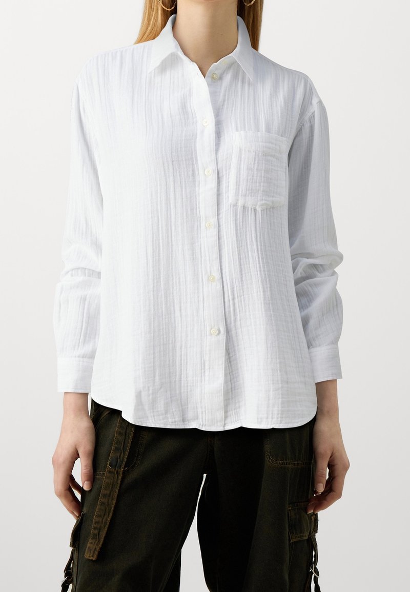 Chemise blanche à manches longues avec boutons, confectionnée en tissu texturé, dotée d'une poche poitrine et d'un ourlet incurvé.