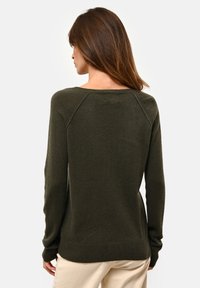 Žena s dlouhými hnědými vlasy, oblečená v tmavě olivově zeleném svetru s dlouhým rukávem a světle béžových kalhotách, zobrazená zezadu.