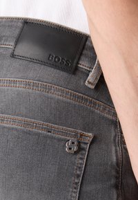 Jeans in denim grigio con etichetta in pelle nera con logo "BOSS" in rilievo. Include cuciture arancioni e un accento in metallo con logo sulla tasca posteriore.