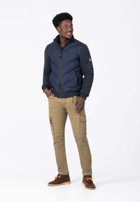 Marine blauwe gecapitonneerde jas met rits, gebreide mouwen; kaki cargo broek met zakken; bruine laarzen met oranje veters. Man staat casual en lacht.