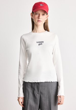 Chemise blanche à manches longues avec un logo brodé "tommy girl", associée à un pantalon gris à rayures. Casquette rouge portée sur la tête.