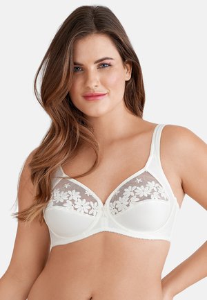 Frau mit langen braunen Haaren, die einen weißen BH mit floralen Spitzendetails auf den Körbchen trägt, leicht lächelnd vor einem einfarbigen Hintergrund.