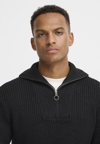 Schwarzer Strickpullover mit hohem Kragen, halbem Reißverschluss und strukturiertem Muster. Verfügt über einen runden Reißverschlussanhänger und eine weiße Innenschicht.