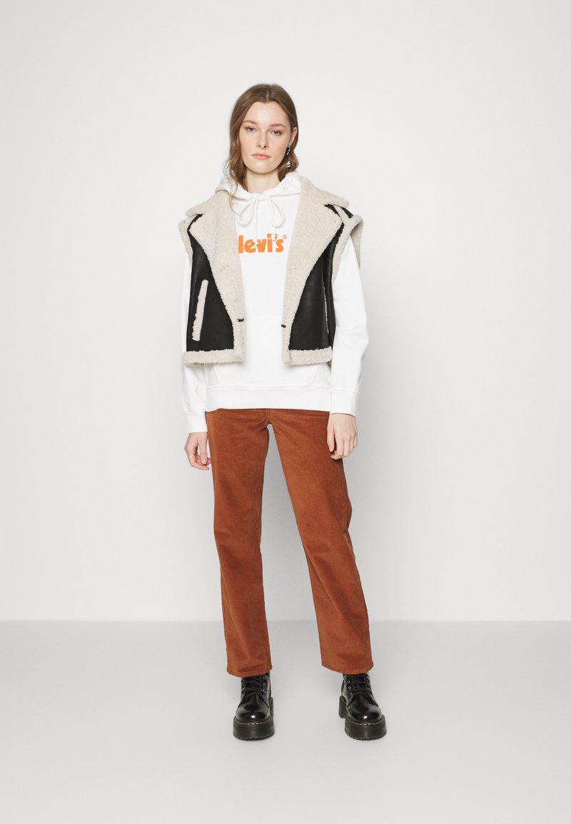 Schwarwest und cremefarbene ärmellose Weste über einem weißen Hoodie mit orangem Logo, kombiniert mit rostfarbenen Cordhosen und schwarzen Schnürstiefeln.