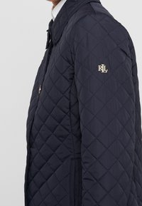 Marinblå, kviltad jacka med ribbad krage, sidofickor och en gyllene logotyp på vänster ärm, med ett strukturerat geometriskt mönster.