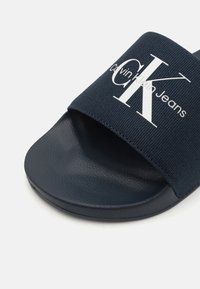 Marine skyddsandal med ett texturerat tygband som har vit "Calvin Klein Jeans" logotyp. Slät gummisula med rundad form.