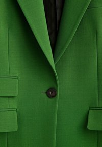 Zielony żakiet wykonany z teksturowanej tkaniny. Wyróżniające się detale to pojedynczy czarny guzik, strukturalne klapy i dwie boczne kieszenie.