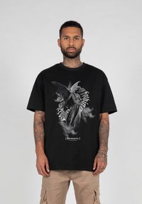 Black oversized t-shirt featuring a detailed angel graphic in grayscale, with white text below and short sleeves. Fabric appears cotton-rich.
