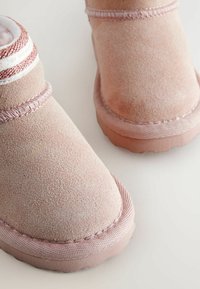 Izbliza pogled na ružičaste zimske čizme od nubuka s gumenim potplatima i dekorativnim šavovima na bijeloj površini.