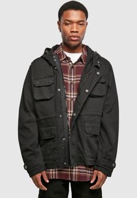 Black hooded jacket with multiple front pockets, button accents, and a textured fabric, worn over a plaid shirt.
