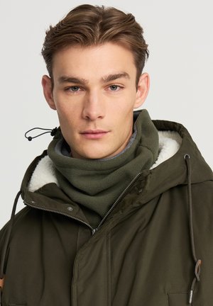 Giovane uomo che indossa un giubbotto invernale verde oliva con cappuccio e un collare in pile coordinato, su uno sfondo semplice.