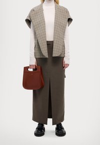 Donna con dolcevita beige, gilet a scacchi, gonna marrone scuro con spacco frontale, scarpe nere, mentre tiene una borsa a tracolla in pelle marrone.