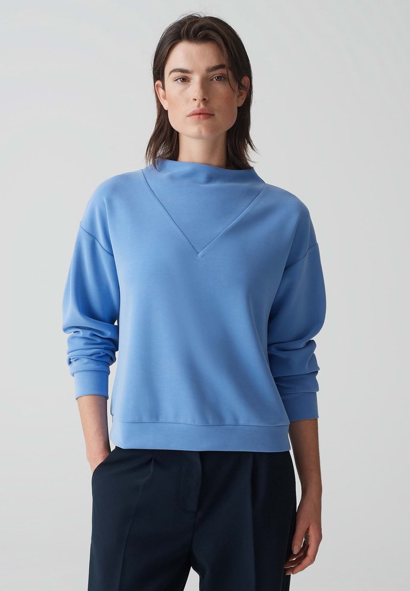 Sweatshirt bleu clair avec un col montant, une coupe décontractée, des manches larges et des poignets côtelés. Texture lisse, design minimal.