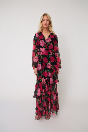 Šviesiaplaukė moteris stovi vilkėdama ilgą juodą suknelę su rožiniais ir raudonais gėlių raštais, plonais ilgomis rankovėmis ir raukinuotais sluoksniais.