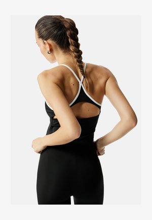 Vrouw met gevlochten haar, die een zwart hemdje met witte bandjes draagt, van achteren gezien, handen in de zij tegen een witte achtergrond.