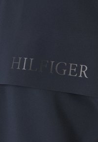 Mørkeblå stof med en glat tekstur, der har et metallisk sølvfarvet "HILFIGER" logo trykt fremtrædende hen over midten.