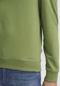 Groene longsleeve sweatshirt met een gestructureerde brei, ribgebreide manchetten en een zijlabel. Gecombineerd met blauwe spijkerbroeken, met de nadruk op de stofdetails.