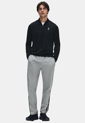 Giovane uomo che indossa un top nero a maniche lunghe con zip e pantaloni grigio chiaro con scarpe da ginnastica nere, in piedi con le mani nelle tasche.