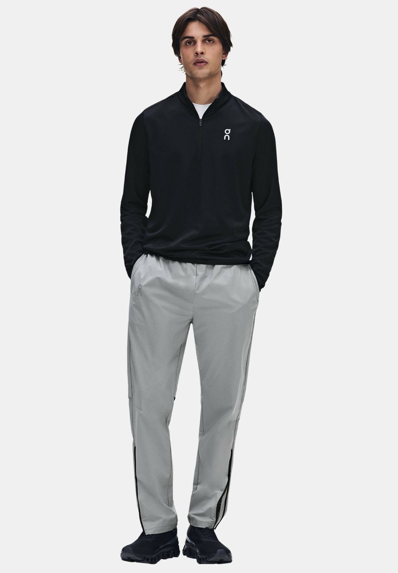 Jeune homme portant un haut noir zippé à manches longues et un pantalon gris clair avec des chaussures de sport noires, debout les mains dans les poches.