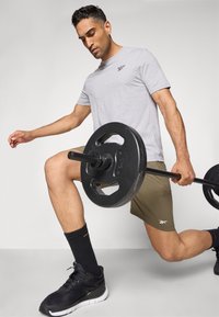 Pelēks sporta krekliņš, olīvu krāsas šorti, melni sporta apavi un zeķes. Tur melnas svara plāksnes ar koncentrētu stāju fitnesa vidē.