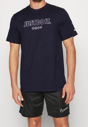 Homme portant un t-shirt bleu marine avec le texte "JUST DO IT." et un short noir Nike avec un logo Swoosh blanc.