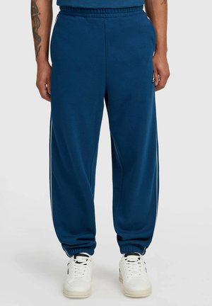 Man wearing blue sweatpants with elastic cuffs and white sneakers, standing against a plain light gray background.