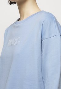 Světle modrý svetr s kulatým výstřihem, s bílým vyšitým logem "HUGO" na hrudi a vroubkovanými manžetami. Jemná textura a pohodlný střih.