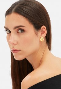 Złote kolczyki wkręty o teksturowanej powierzchni i abstrakcyjnym kształcie, noszone na uchu modelki, podkreślające ich refleksyjną jakość i wzornictwo.