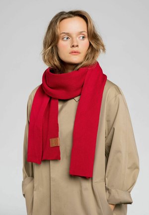 Czerwona, dziergana szalik o ryżowym wzorze, wyeksponowany owinięty wokół szyi. Skórzana metka na jednym końcu; noszony na beżowym płaszczu.