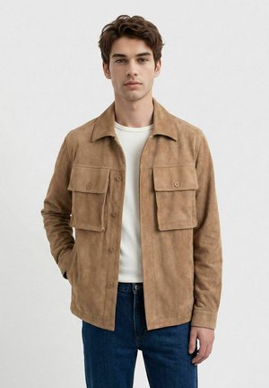 Ung mann iført en beige semsket jakke med knappede klaffelommer over en hvit skjorte og blå jeans, stående mot en ensfarget bakgrunn.