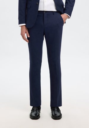 Homme portant un pantalon de costume bleu marine, une veste assortie, une chemise blanche et des chaussures habillées noires, une main dans la poche, sur un fond clair uni.
