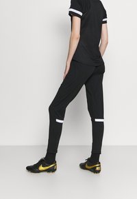 Schwarzes Sportoutfit mit einem schwarzen Oberteil mit weißen gestreiften Ärmeln und schwarzen Hosen mit weißen Akzenten; getragen mit schwarz-goldenen Turnschuhen.