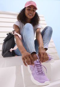 Lila Netzsneaker mit einer weißen Sohle, ausgestattet mit einer Schnürung und einem auffälligen Logo, kombiniert mit hellblauen Jeans und einem grauen Rucksack.