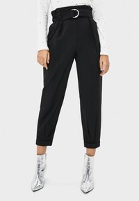 Hoch taillierte schwarze Hose mit einem Gurtbund, gefältelter Vorderseite, schmal zulaufendem Bein und umgeschlagenen Säumen, kombiniert mit glänzenden silbernen Knöchelstiefeln.