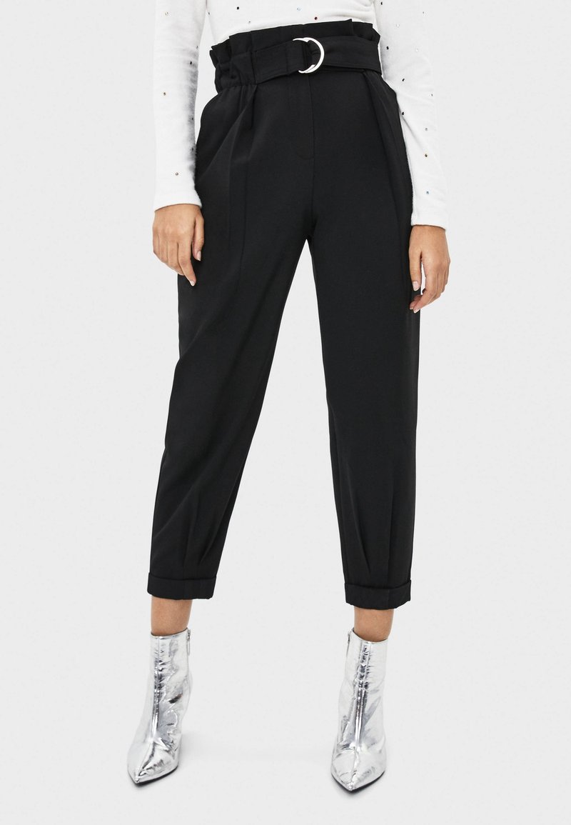 Hoch taillierte schwarze Hose mit einem Gurtbund, gefältelter Vorderseite, schmal zulaufendem Bein und umgeschlagenen Säumen, kombiniert mit glänzenden silbernen Knöchelstiefeln.