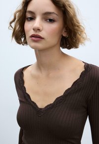 Brun ribstrikket top med lange ærmer og skålsyet blonderubrik ved halsen. Glat tekstur med en tætsiddende silhuet, der fremhæver subtile variationer i hudtonen.