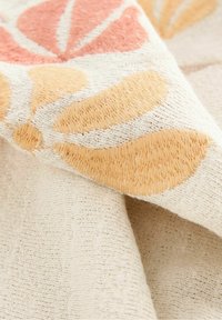 Kremowa tkanina z teksturową, żółtą i różową haftowaną florystyką w miękkich, nakładających się fałdach.