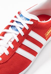 Röd mocka sneaker med vita läderränder, rund tå och platta vita snören. Har en blå och vit logotyp på tungan. Röd inredning.