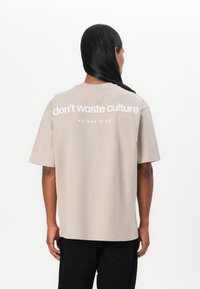 Beige T-shirt lavet af bomuld, med korte ærmer og runde udskæring. Bagsiden har design med hvid tekst: "spild ikke kultur MAISON D'ART."