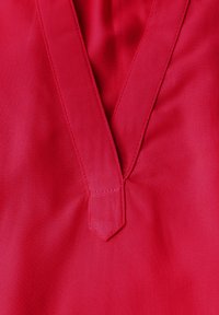 Tessuto rosso con una consistenza liscia presenta un collo a V e un dettaglio appuntito al centro. Le cuciture sono visibili lungo i bordi.