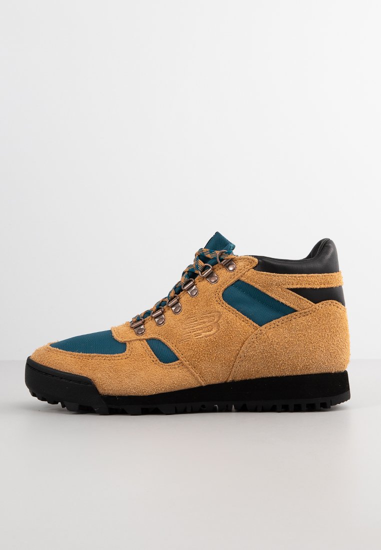 Tan suède en teal stof wandelschoen met zwarte zool en metalen oogjes voor de veters, zijkant bekeken op een plain witte achtergrond.