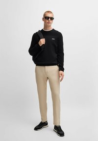 Felpa nera con logo bianco, pantaloni beige slim-fit e sneakers nere. Il modello tiene uno zaino nero e indossa occhiali da sole scuri.