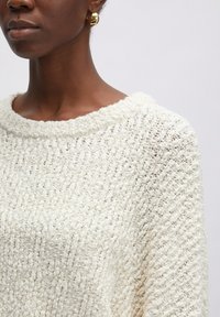 Strickpullover in Off-White mit einem strukturierten, locker gewebten Muster. Verfügt über einen weiten Ausschnitt und eine lässige Passform. Goldene Steckerohrringe.