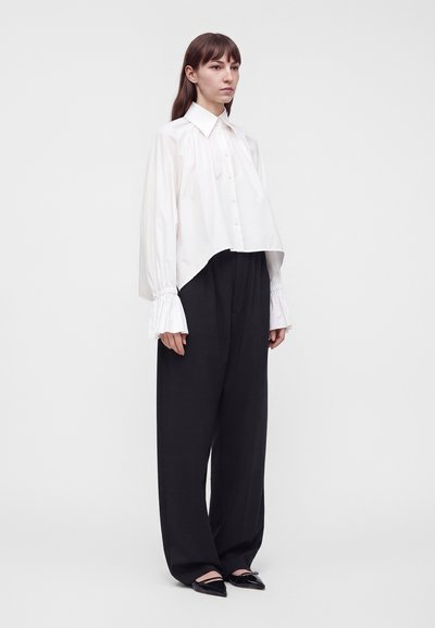 Femme portant une blouse blanche ample boutonnée avec des poignets évasés, un pantalon noir à jambes larges et des chaussures noires pointues, debout devant un fond blanc.