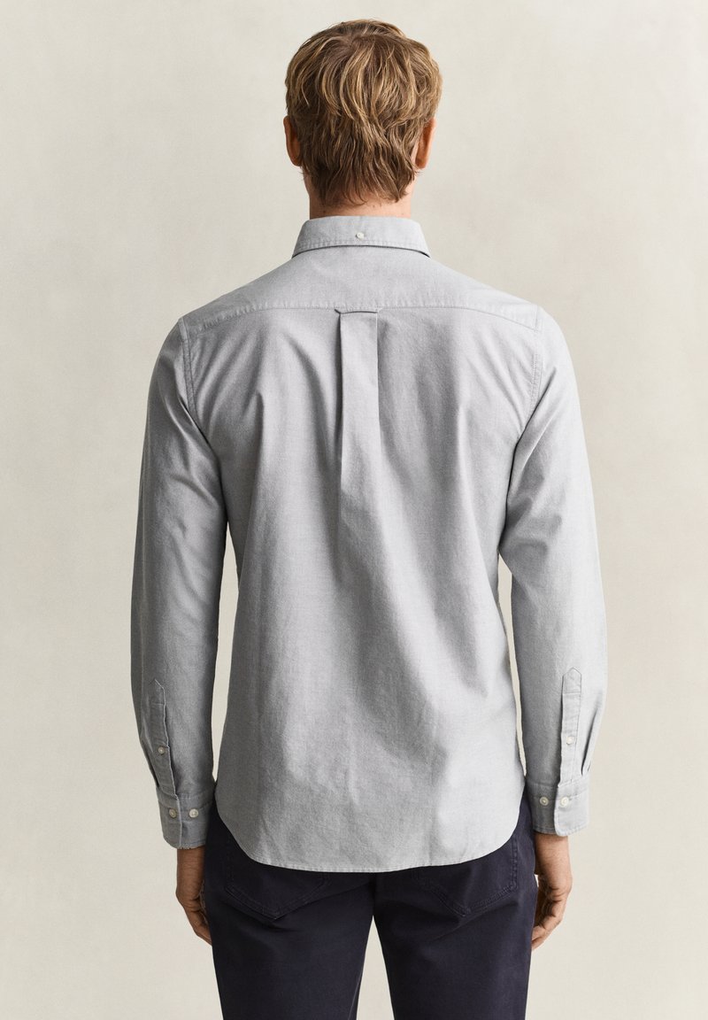 Homme aux cheveux blonds courts portant une chemise gris clair à manches longues boutonnée et un pantalon foncé, dos à la caméra.