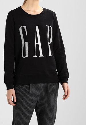 Sweatshirt noir à col rond avec de grandes lettres argentées "GAP". Porté avec un pantalon à rayures gris. Matière en coton doux.