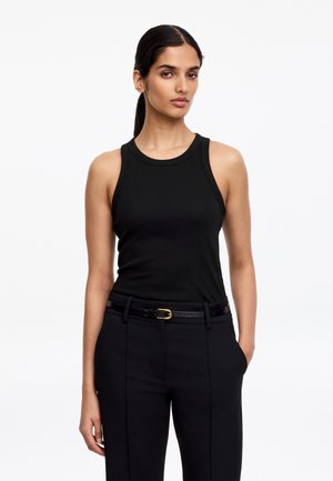 Femme aux cheveux foncés attachés en arrière, portant un haut noir sans manches côtelé, un pantalon noir et une ceinture noire avec une boucle dorée, debout devant un fond blanc.