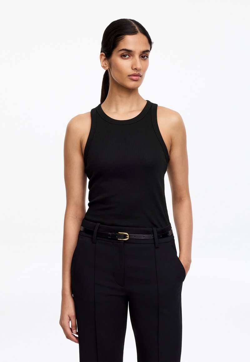 Femme aux cheveux foncés attachés en arrière, portant un haut noir sans manches côtelé, un pantalon noir et une ceinture noire avec une boucle dorée, debout devant un fond blanc.